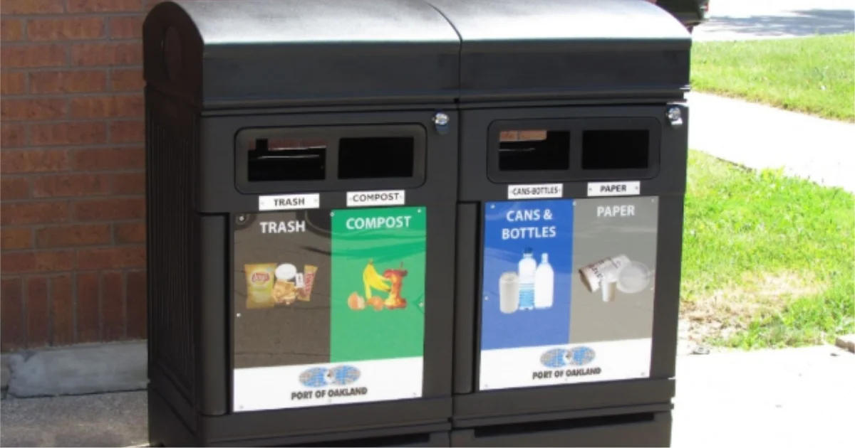 Outdoor waste and recycling station with labeled bins for trash, compost, cans and bottles, and paper, located on a sidewalk.